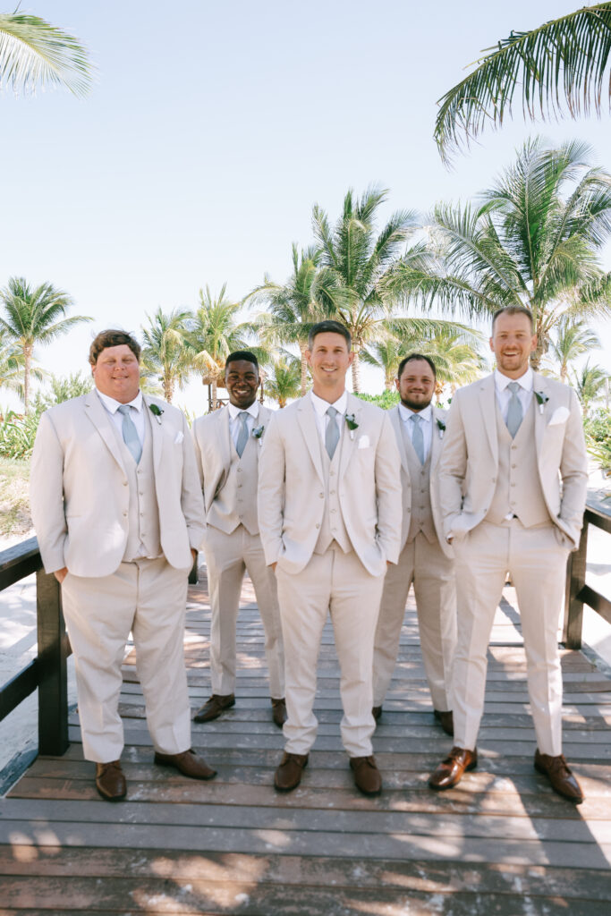 Groom and Groomsmen in beige tuxedos with blue ties standing in a staggered formation on a wooden boardwalk with palm trees in the background