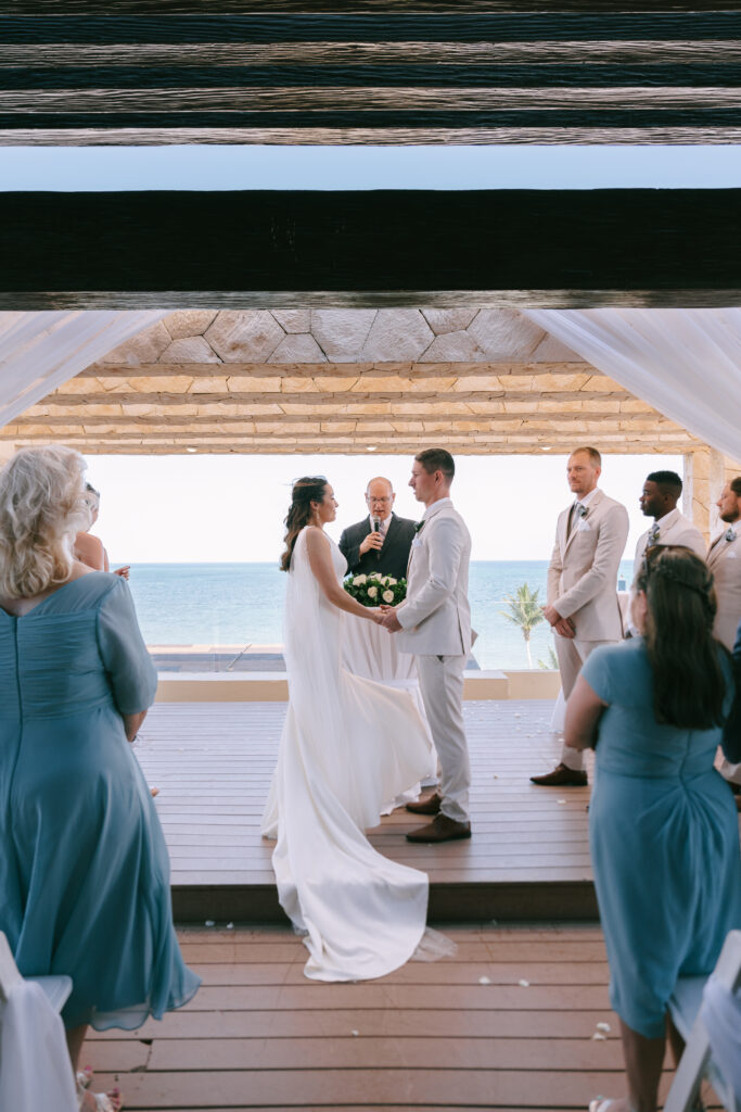 Bride and Groom standing at the alter holding hands while wedding ceremony is occurring. Family and friends in blue stand to the left and right of the couple.