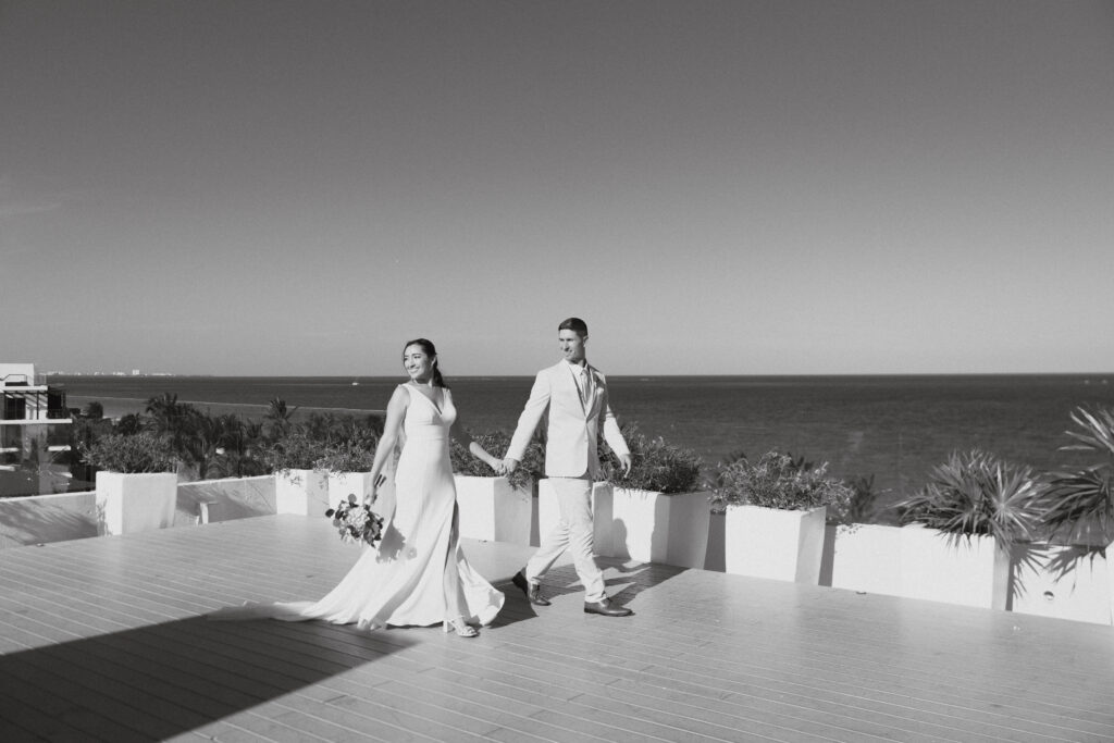 Bride and groom walk hand in hand on a patio with the sea behind them at the Royalton Riviera Cancun resort