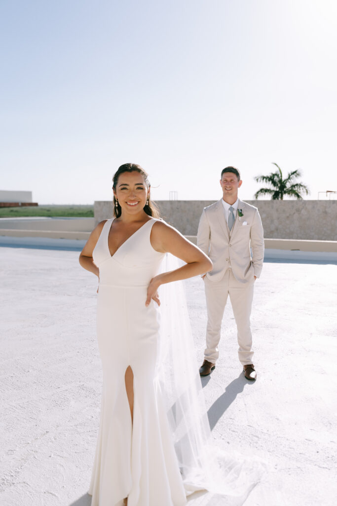 Bride in wedding dress stands smiling with hands on her hips. Groom in background stands gazing at Bride with hands in pockets.