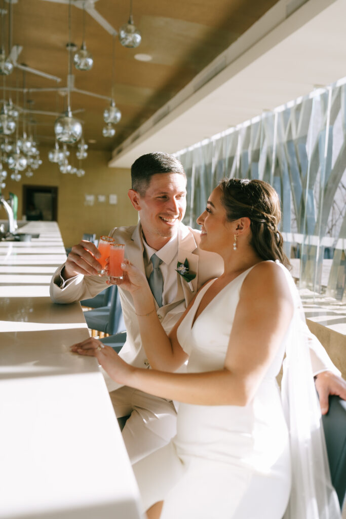 Bride and Groom touching glasses and gazing at one another seated at a white bar.