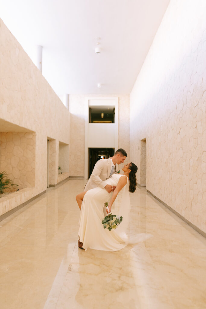 Groom dipping Bride to the right in a long white and beige marbled hallway.