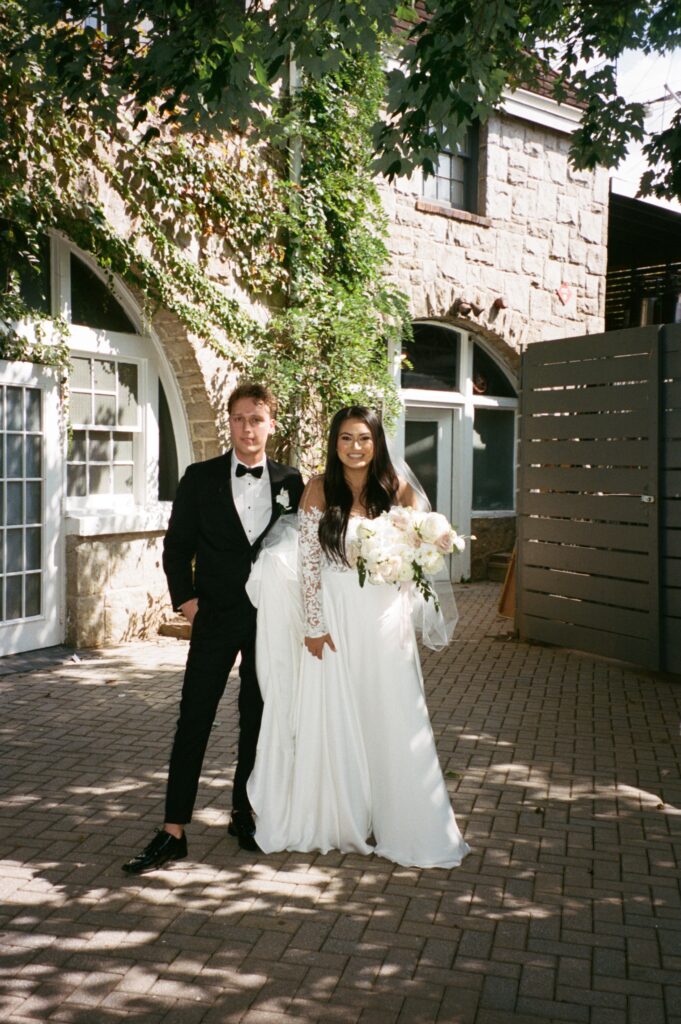 Groom in a black tuxedo stands to the left of a bride in a long sleeve lace gown. A stone building covered with ivy sits in the background.