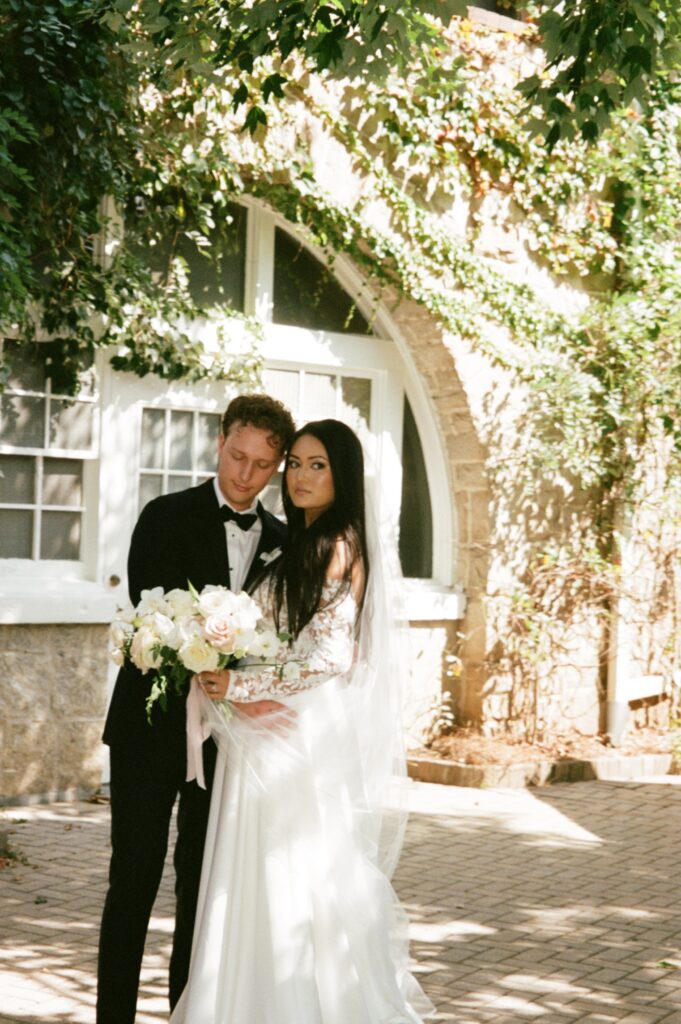 Bride and groom stand with heads touching in a brick courtyard with an ivy covered stone building in the background.