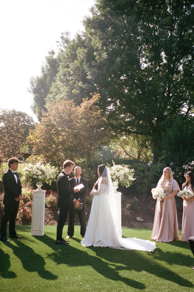 Bride and Groom stand face to face on a green lawn in front of the officiant. Trees of various sizes cover the background.