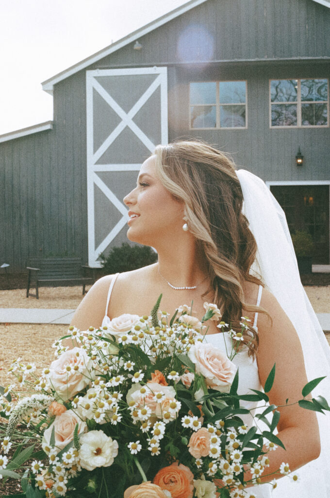 Bride gazing over her shoulder, with a bouquet of colorful roses and other florals, and  a large grey barn in the background