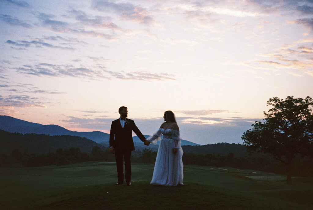 Bride and Groom stand with hands held gazing at each other facing the viewer. The picture is dim and the sunset glows orange and yellow in the background.