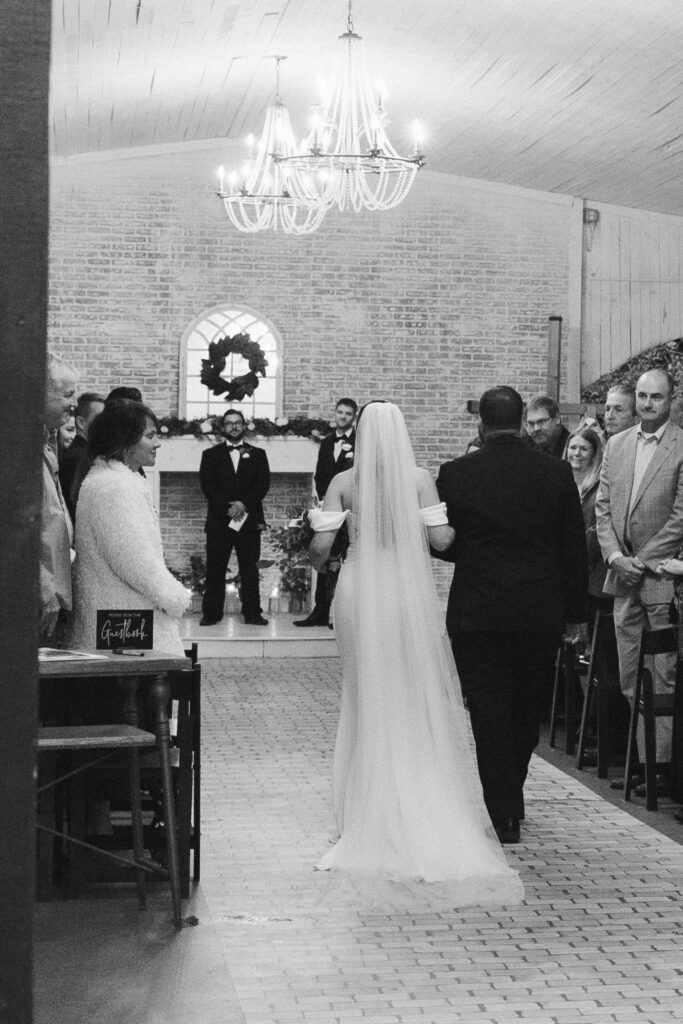 Black and white image of father walking bride down the stone aisle, away from the viewer, toward the groom and officiant standing in front of a brick wall. Family and friends stand on either side with a chandelier overhead 