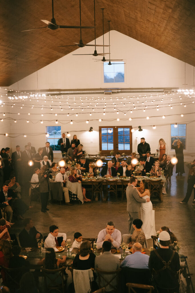 Bride and Groom share a first dance surrounded by family and friends sitting at long rectangular tables in a white-walled barn with bistro lights overhead.