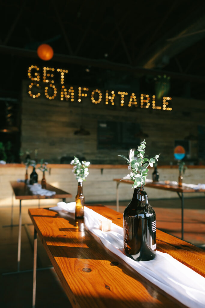 Electric sign that says Get Comfortable on a wooden wall at Creature Comforts Brewery. Tables with white runners and greenery in glass beer bottles.