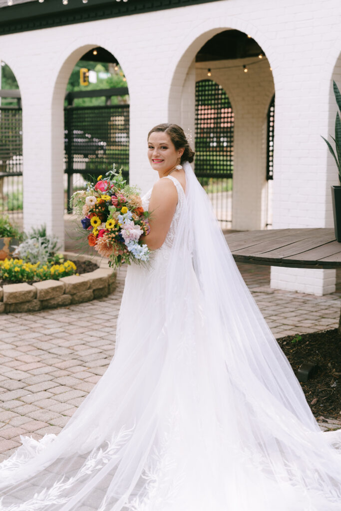 Bride in a white dress with a multicolored flower bouquet gazing over her shoulder at the camera. She stands on a brick courtyard with white curved arches in the background.