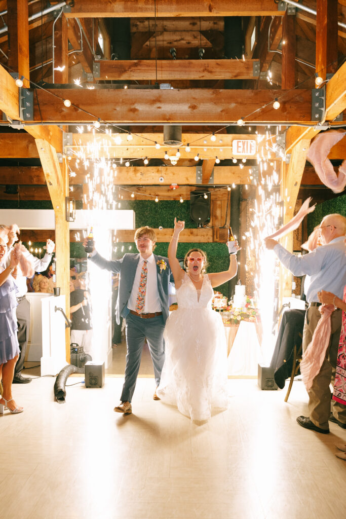 Bride and Groom walking toward the camera with exposed wooden beams overhead, arms raised in excitement. Large sparklers erupt on either side.