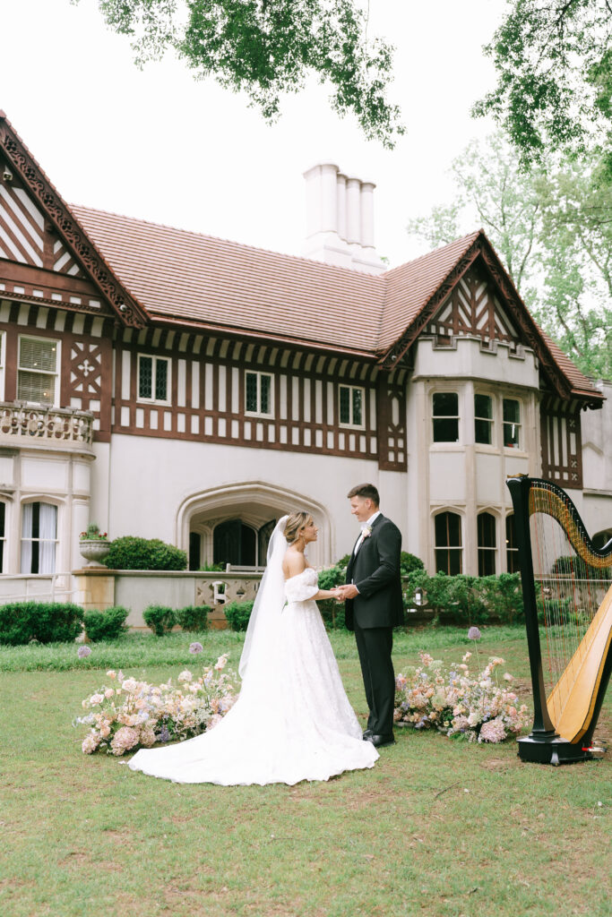 Bride and Groom stand hand in hand gazing at each other on a green lawn with a white brick and wood lattice, European style manor in the background.