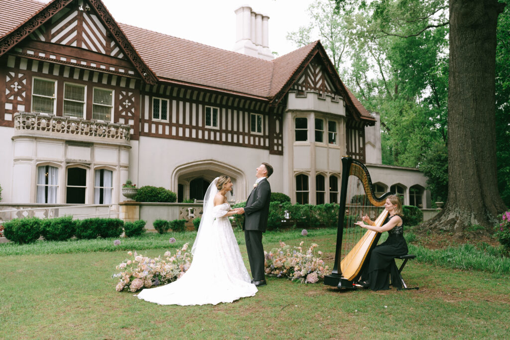 Bride and Groom stand hand in hand laughing on a green lawn with a white brick and wood lattice, European style manor in the background.