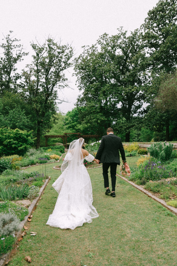 Bride and Groom walk away from the viewer towards a bright green garden surrounded by trees.