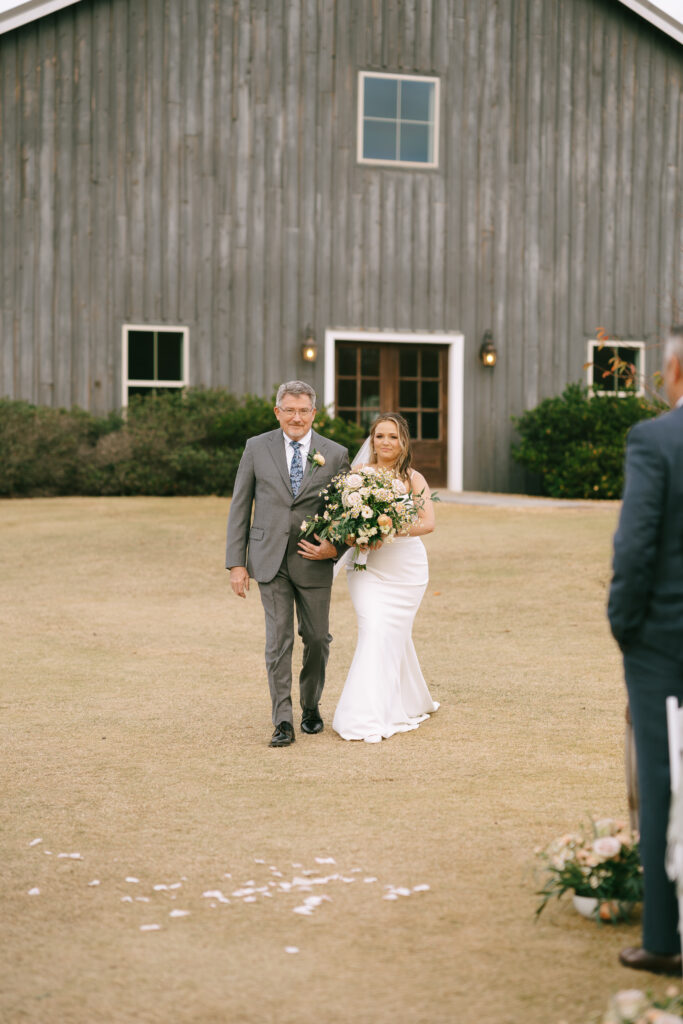 Father of the Bride walking arm in arm with Bride down an outdoor aisle, a large grey colored barn in the background.