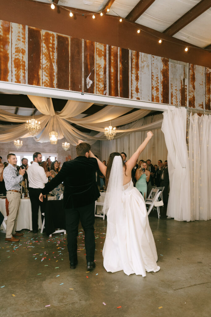 Bride and Groom facing away from the camera entering the reception filled with family and friends. Concrete floors and a colorful wooden mosaic frame the couple.