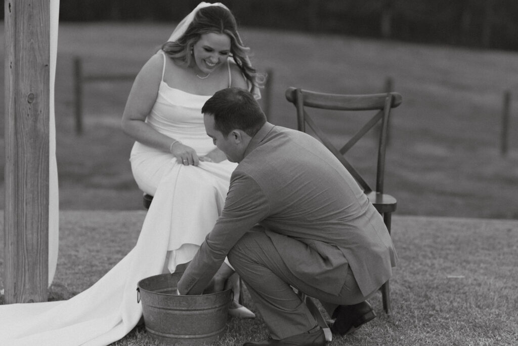 Black and white photo of groom kneeling to wash Bride's foot in a metal bucket. Bride smiles and gazes down at him.