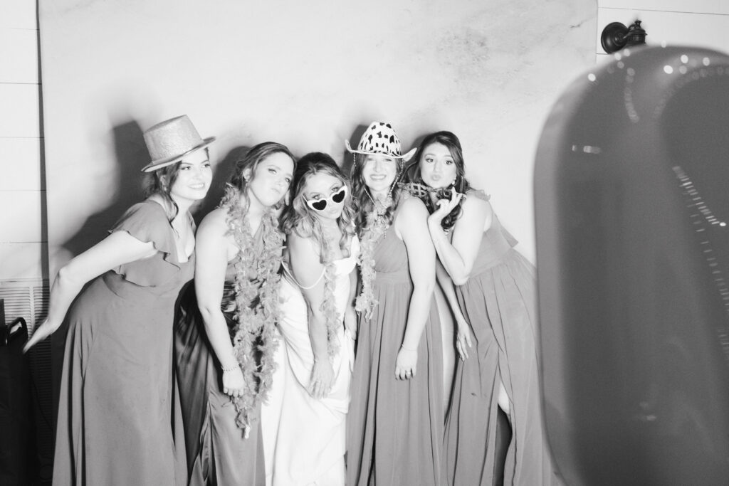 Black and white photo of Bride and Bridesmaids posing in hats and boas for a photobooth.