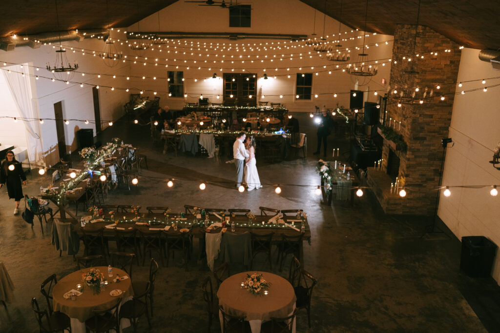 Bride and Groom dancing in the middle of a warmly lit barn at Grant Hill Farms with a stone fireplace and rectangular tables around the perimeter with bistro lights strung overhead.