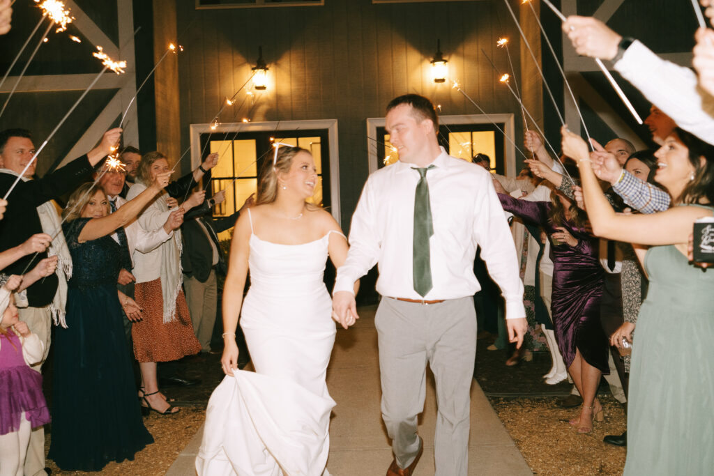 Bride and Groom walking hand in hand, gazing at each other and smiling under an archway made of glowing sparklers held by family and friends.