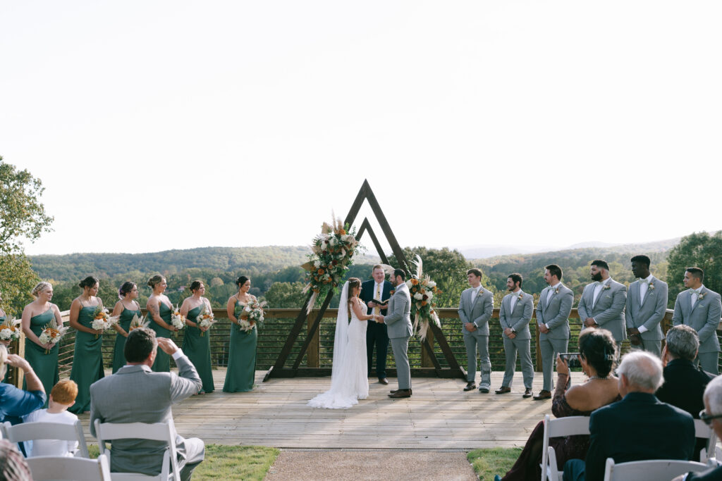 Bride and Groom stand face to face, holding hands outside, in front of a floral covered triangular structure with officiant.