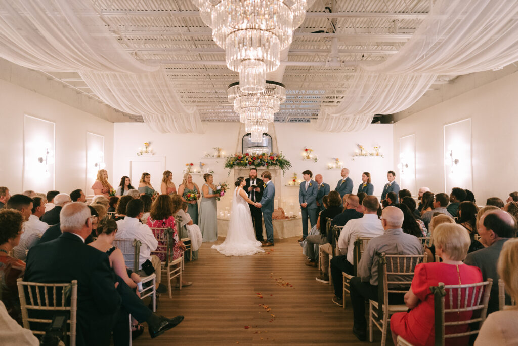 Bride and Groom stand hand in hand on a wood floor in front of a white brick fireplace with large circular chandeliers above and family and friends on either side. 