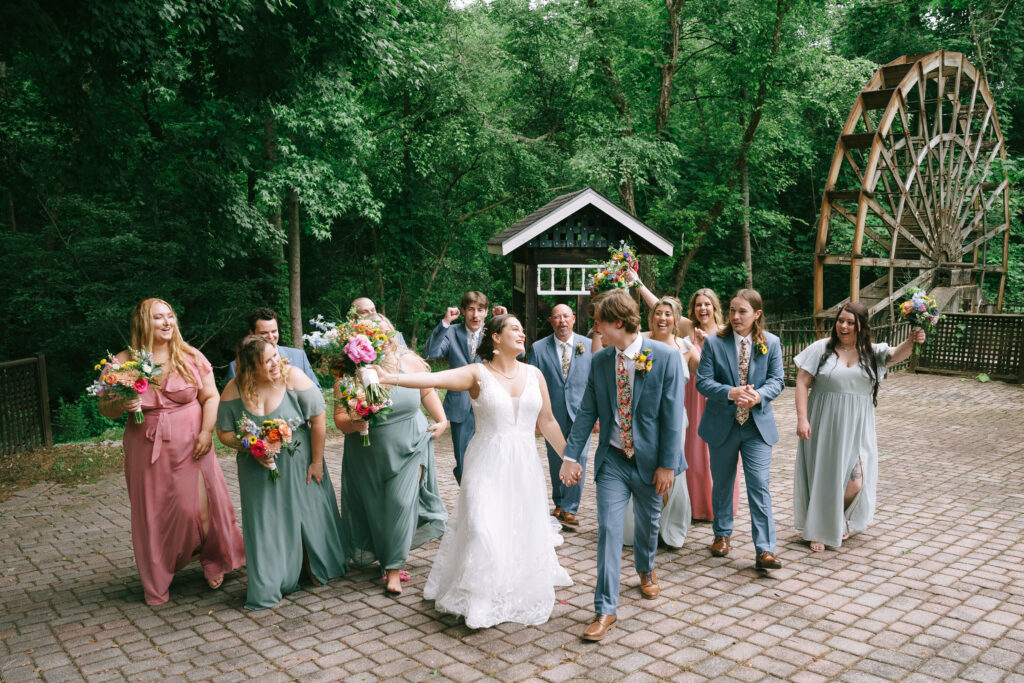 Bride and Groom walk hand in hand gazing at each other with bridesmaids and groomsmen on either side looking toward the bride and groom. Green trees and an old wooden water wheel dominate the background.