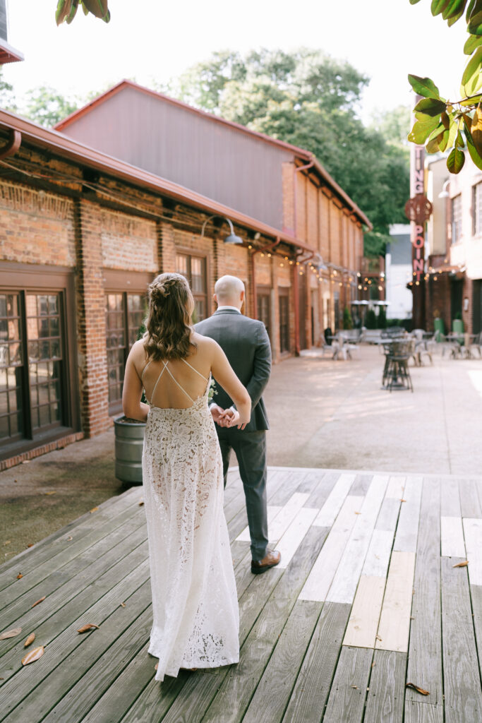 Bride walks towards groom, both facing away from the viewer, on a wooden patio with a brick building on their left.