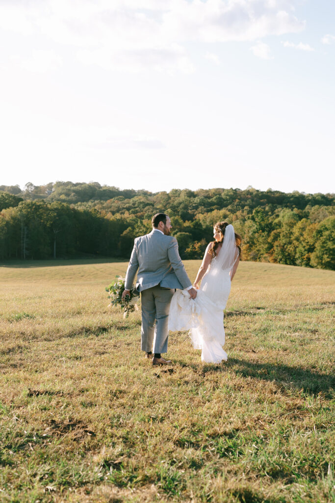 Bride and Groom walk hand in hand away from the viewer in an open field with golden trees in the background.