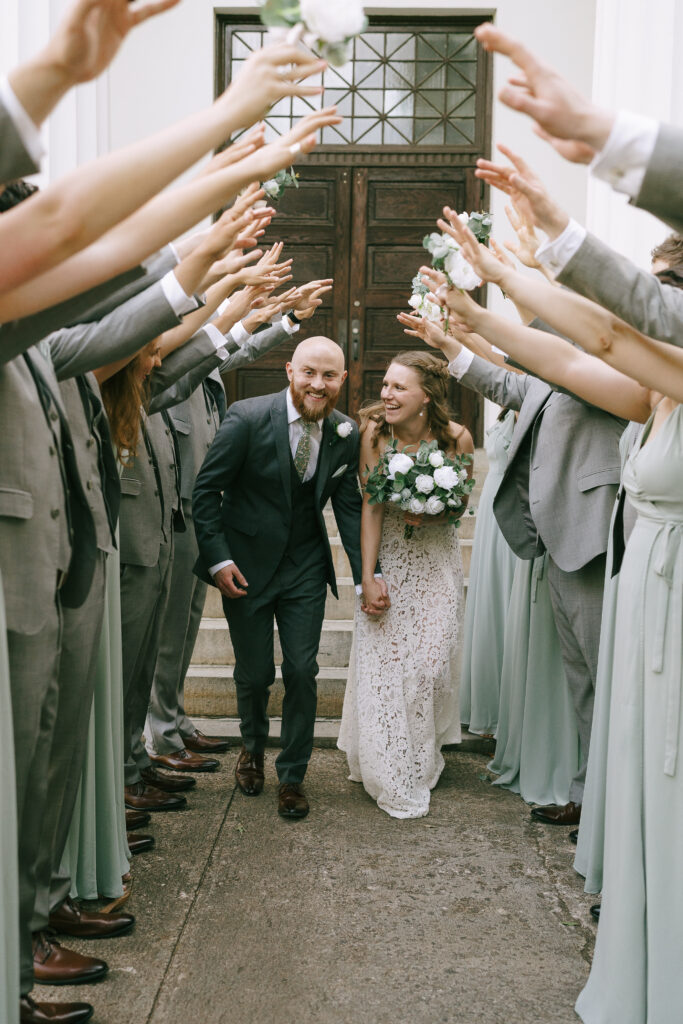 Bride and groom smile, walking hunched towards the viewer under raised arms of bridesmaids and groomsmen.