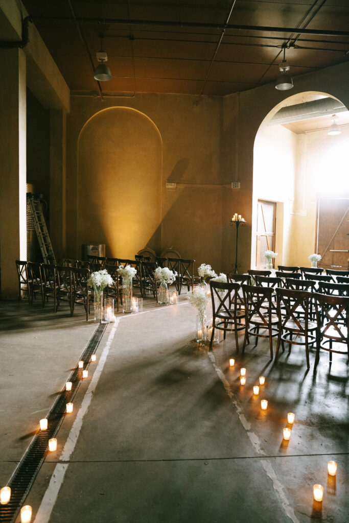 A wedding aisle is bordered by warm, glowing candles, bending around wooden cross-back chairs to a sunlit archway and wooden doors.