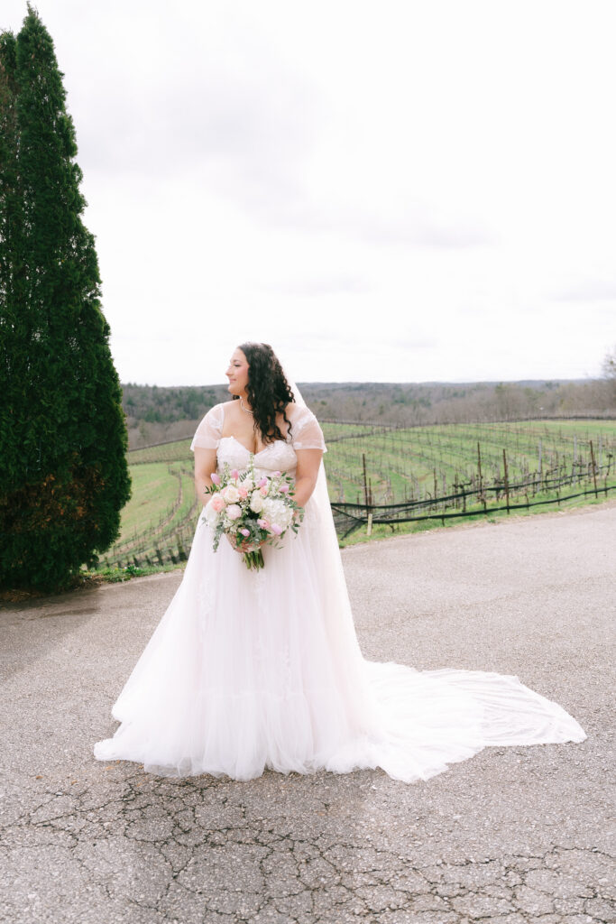 Bride stands facing the camera with her head turned to the left, holding a bouquet of white flowers. She stands on a concrete driveway with rows of a vineyard in the backround.