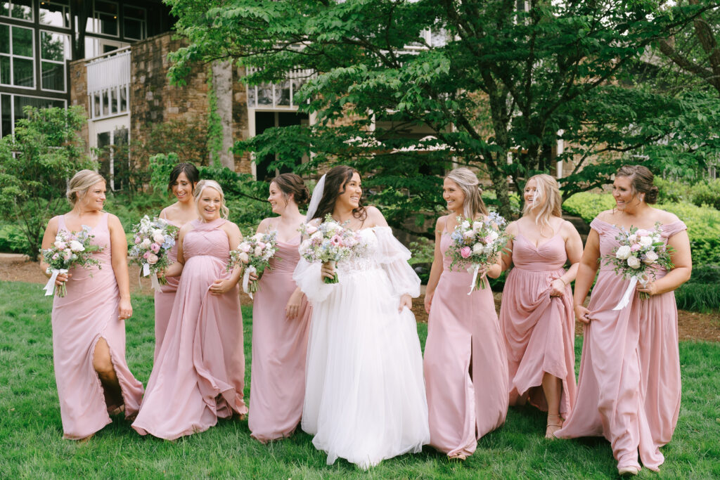 Bride walks with bridesmaids, in pink dresses holding bouquets of pink and white flowers, towards the viewer with green grass and trees. An old brick building sits in the background.