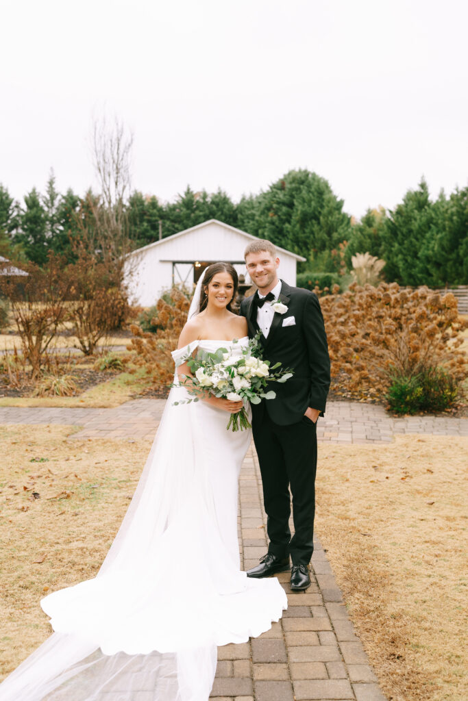 Bride and groom stand side by side on a brick path facing the viewer and smiling. A white bar sits in the background, and golden grass and bushes are between the couple and the barn.