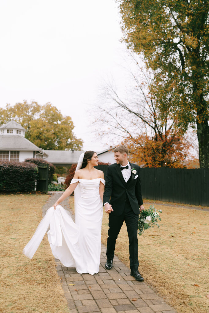 A Bride and groom gaze at each other, holding hands and walking toward the viewer on a brick path. A white building sits in the back left of the image, and golden grass surrounds the path.