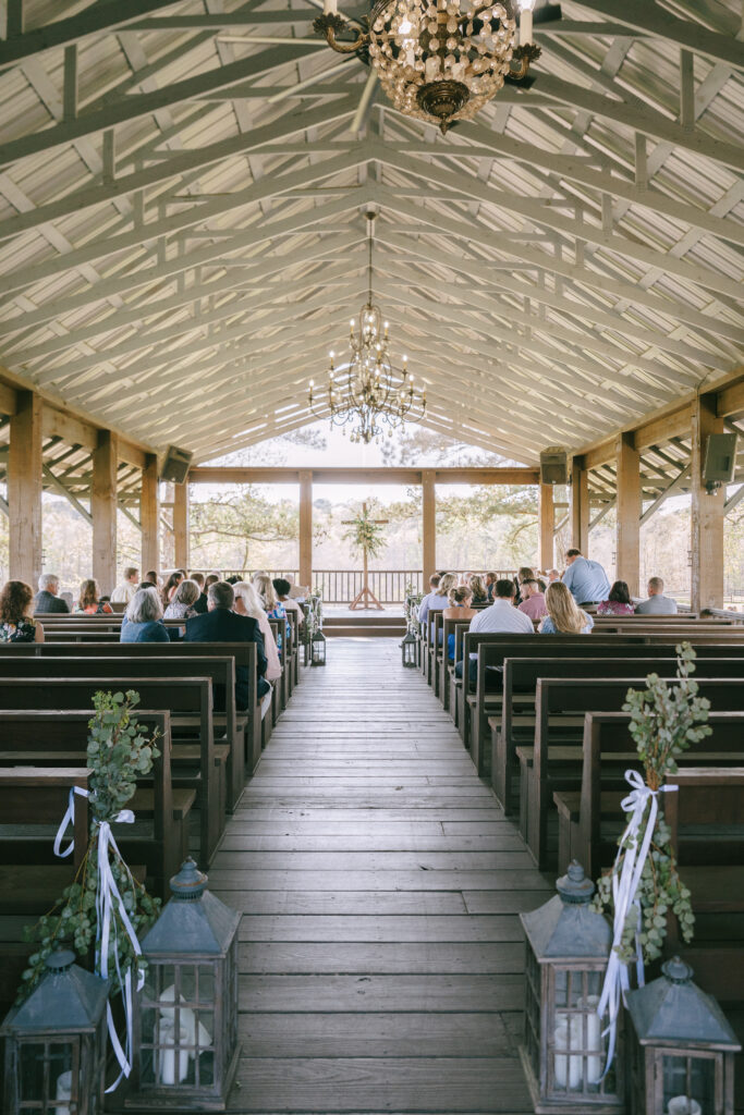 Large windows frame the border of a wooden interior church with wooden pews on either side of the wedding asile.