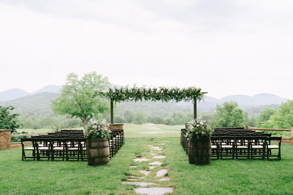 Wooden arbor stands in the background topped with florals with wooden chairs and wooden barrels flanking either side. A stone path creates a wedding aisle surrounded by grass with mountains in the far background.