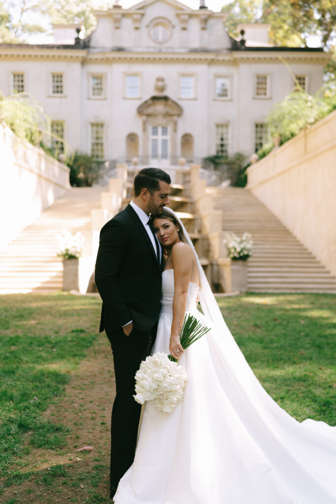 Bride stands with her head on the groom's chest in front of two stone staircases leading to a large, cream colored mansion.