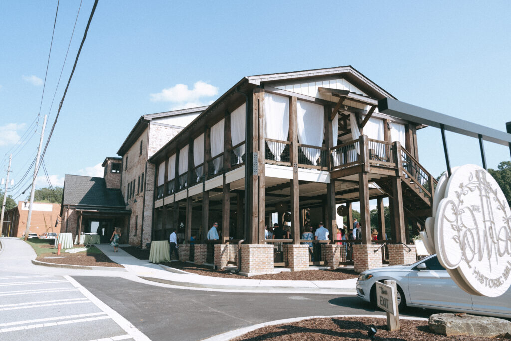 A brick building with wood columns and industrial beams. White drapery appears in the windows of the second story and patrons on the lower patio.