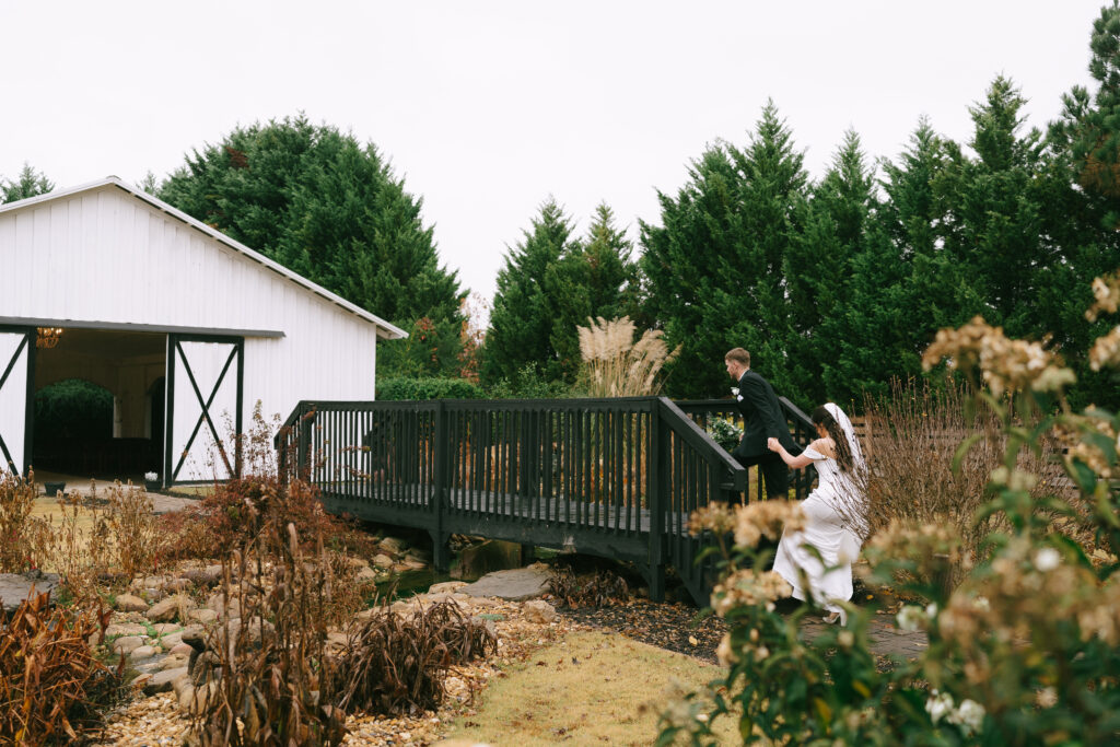 Groom leads bride by the hand up wooden stairs to a bridge above a small waterway filled with river stones. A white bar appears to the left and green trees in the background.