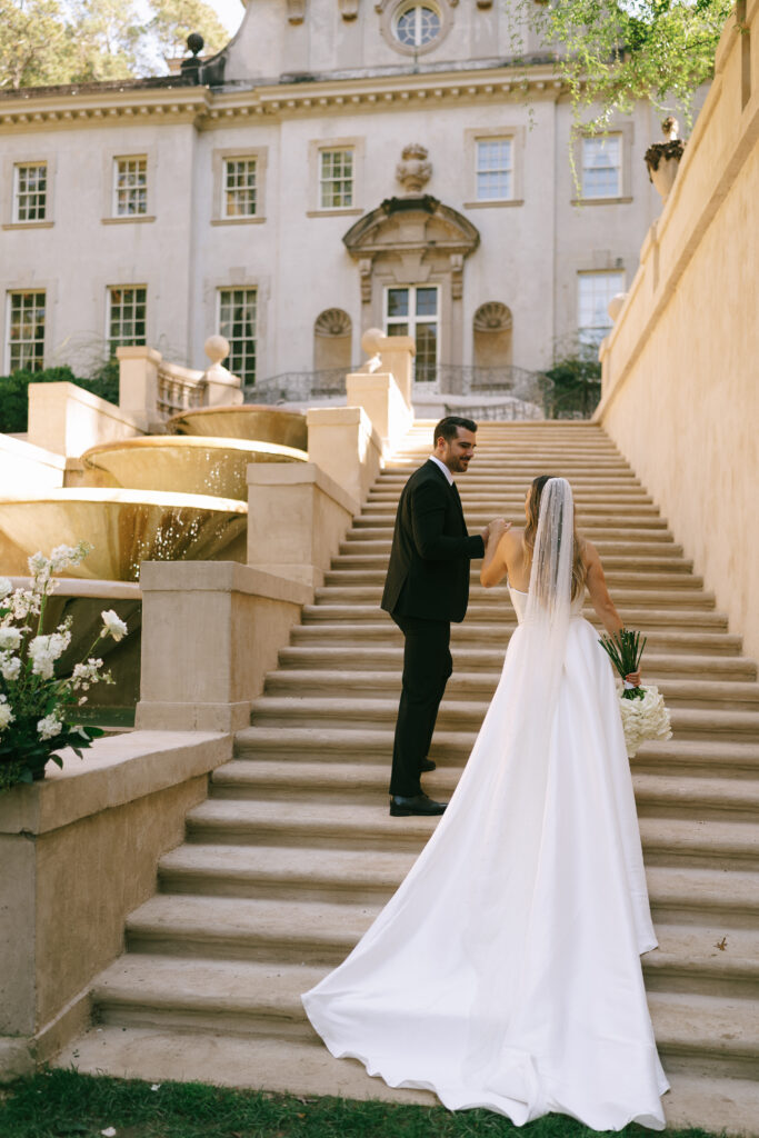 Groom leads bride up a stone staircase with a large, cream colored, two story manor in the background.