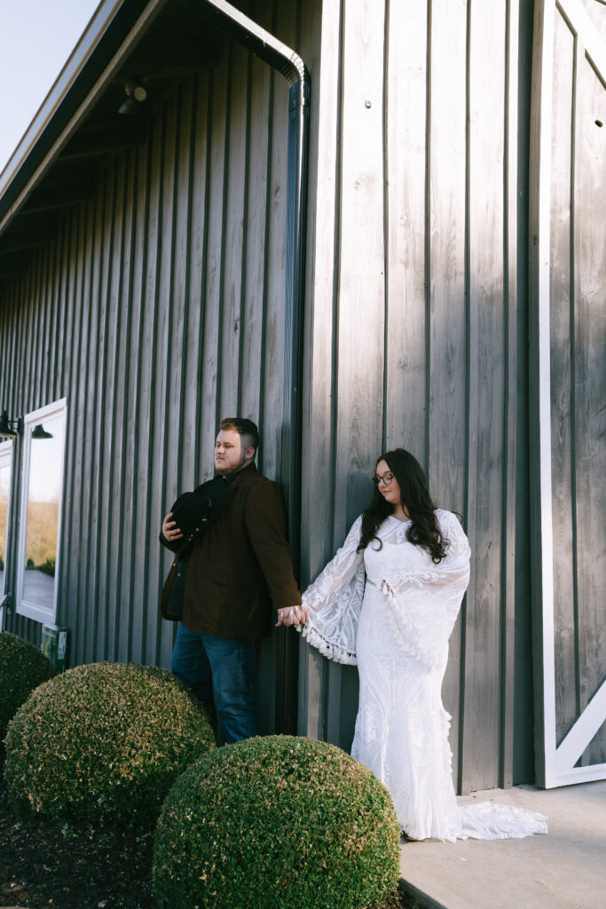 Bride and groom stand around a wooden grey corner holding hands.