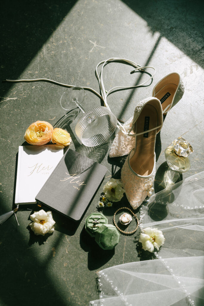 Bridal shoes, florals, vow books reading "his" and "hers", and jewelry arranged on a grey and white marbled concrete floor.