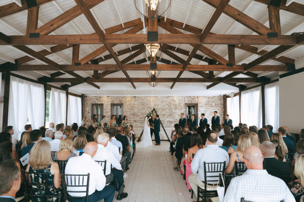 Bride and groom stand hand in hand at the end of a wooden aisle in front of a lime washed brick wall and triangular wooden structure, with family and friends on either side. Exposed wooden beams and metal industrial lighting above.