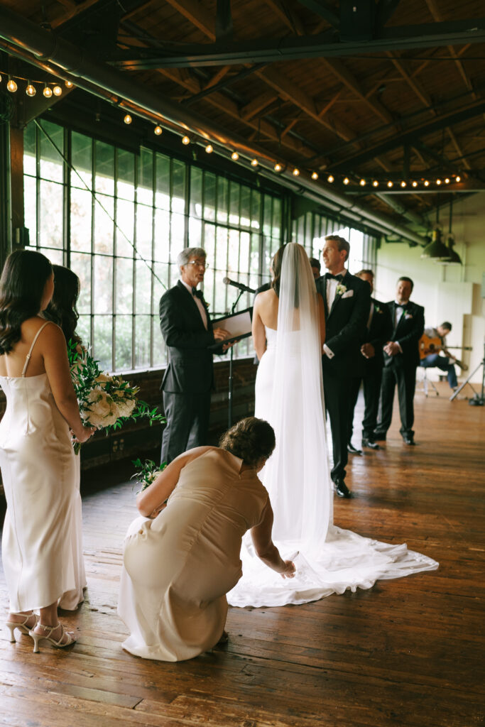 Bridesmaid fixes Bride's train in a wood covered room with rectangular windows.