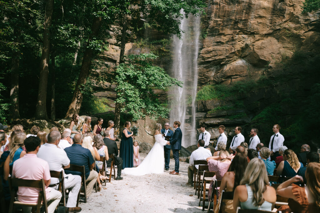 Bride and groom stand hand in hand facing each other, with family and friends seated to either side outside. A large rock face with a waterfall cascading down in the background.