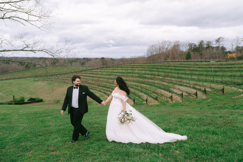 Groom leads Bride by the hand across green grass with vineyard rows in the backround.