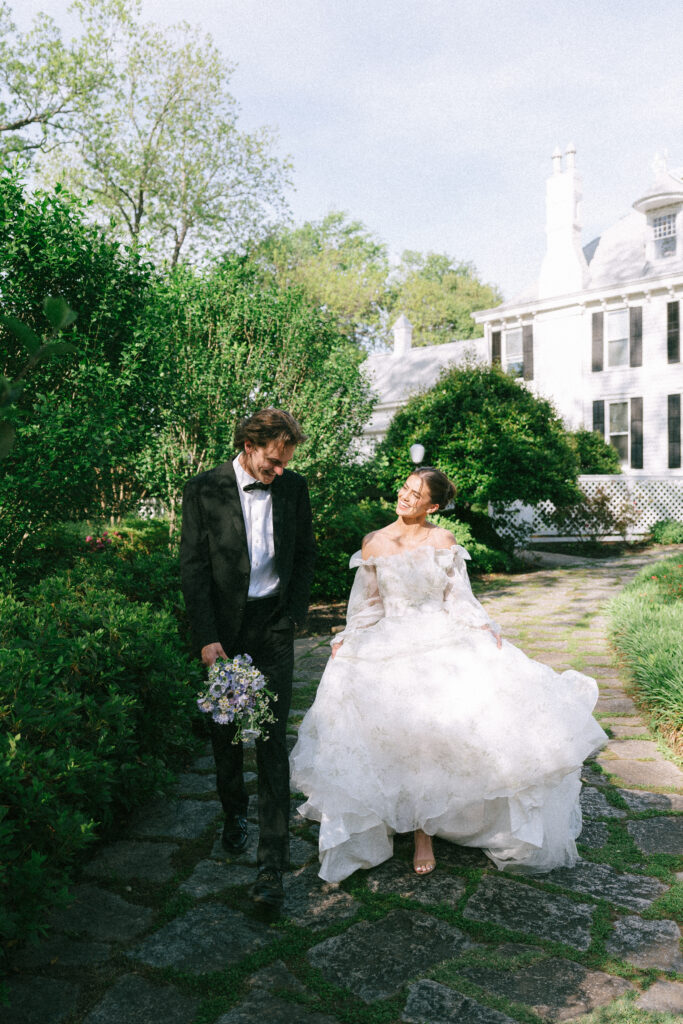 Bride swishes long sleeve gown walking next to groom on a grass filled stone path. A large, white, plantation style manor sits in the background.