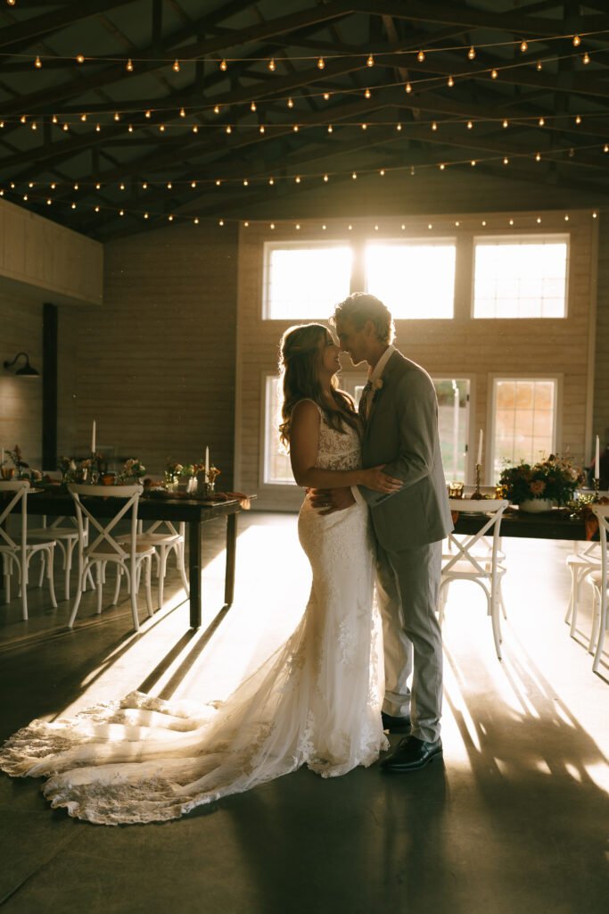 Bride and Groom embrace in a backlit room with bistro lights overhead and sunlight streaming in the background.